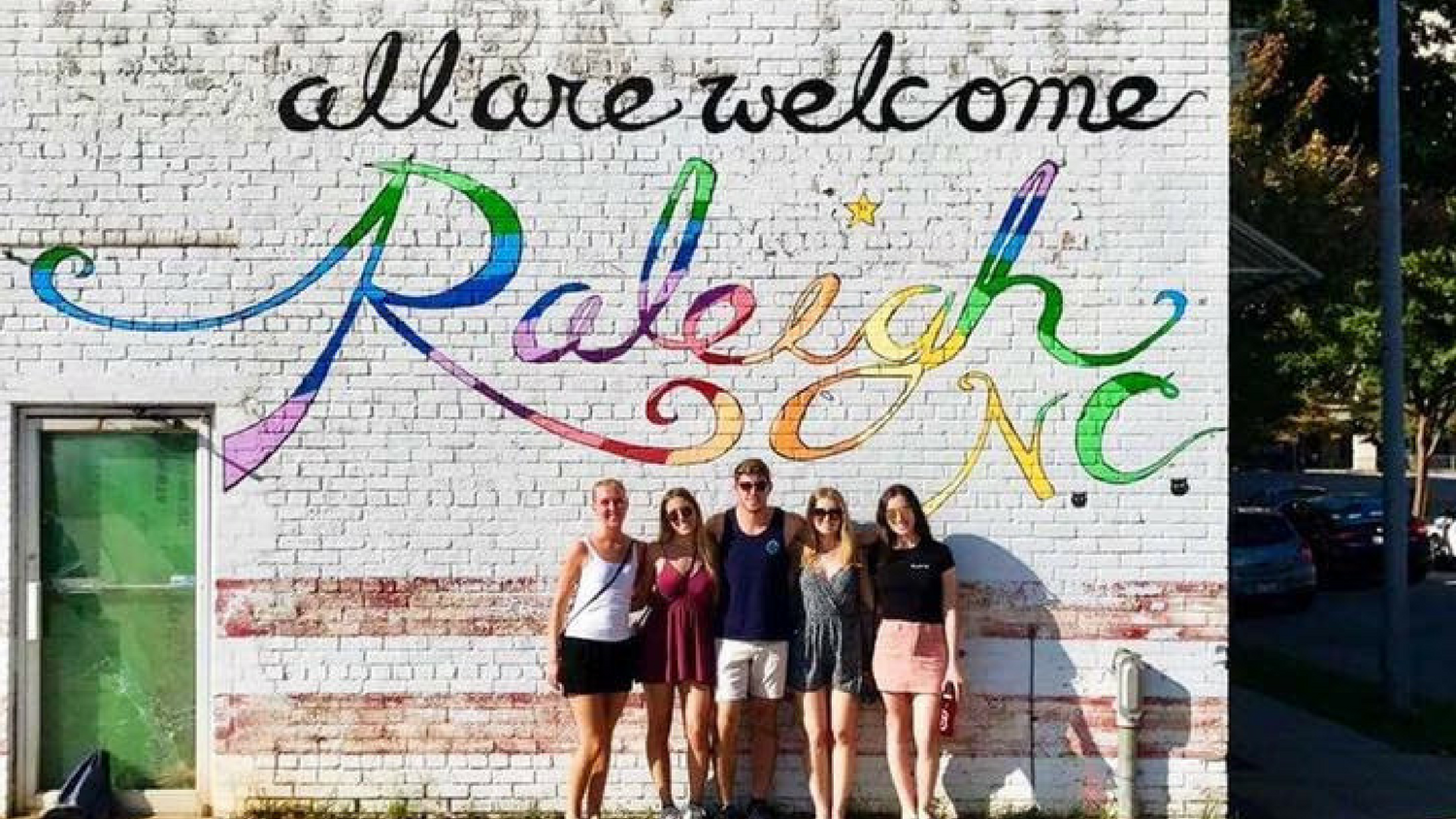 Five students in front of mural on wall painted "all are welcome Raleigh NC" in multicolor letters